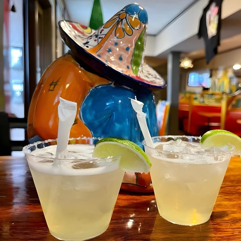 Two margaritas with lime wedges and straws on a wooden table, with a colorful sombrero statue behind.