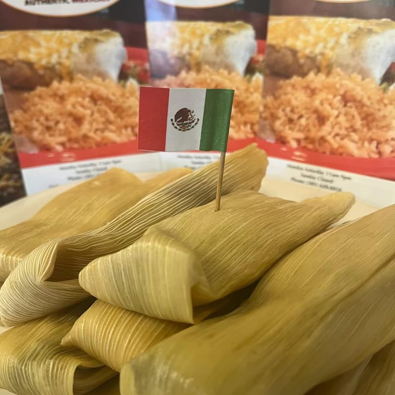 A plate of Cafe Bueno tamales with a small Mexican flag, and menu items in the background.
